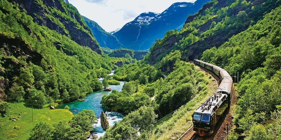 A black train emerges from a tunnel passing through a mountainside, it is surrounded by greenery and next to a bright blue river. 